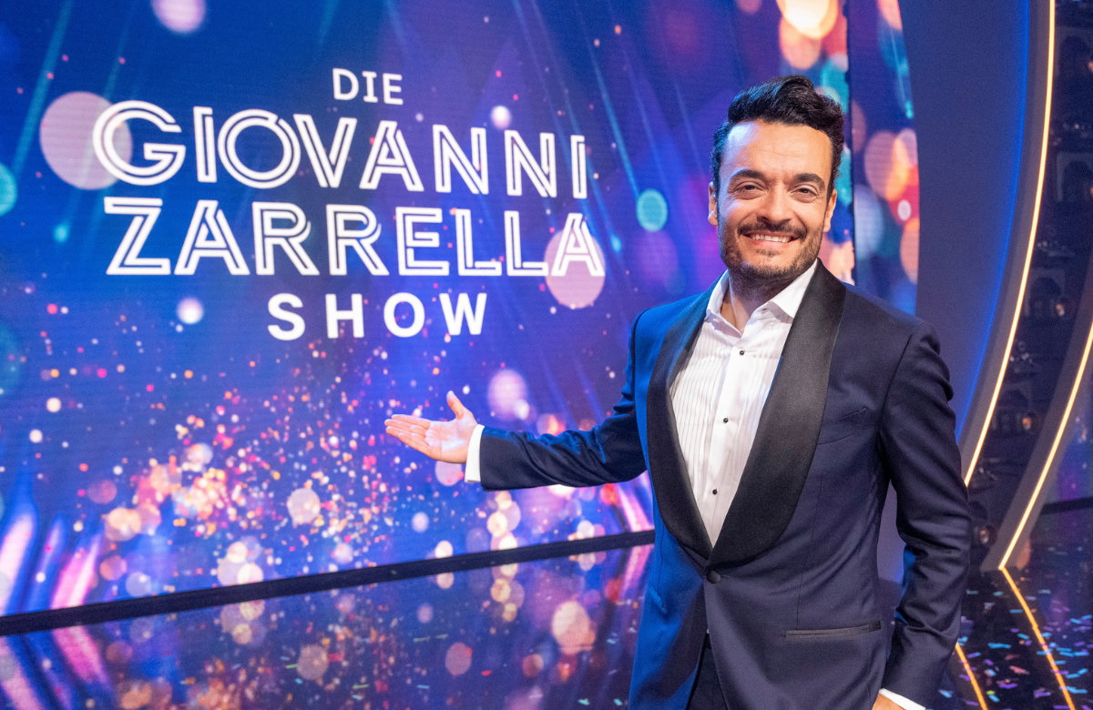 Giovanni Zarrella grinst und zeigt mit ausgestrecktem Arm auf eine Leinwand mit dem Logo der Giovanni Zarrella Show