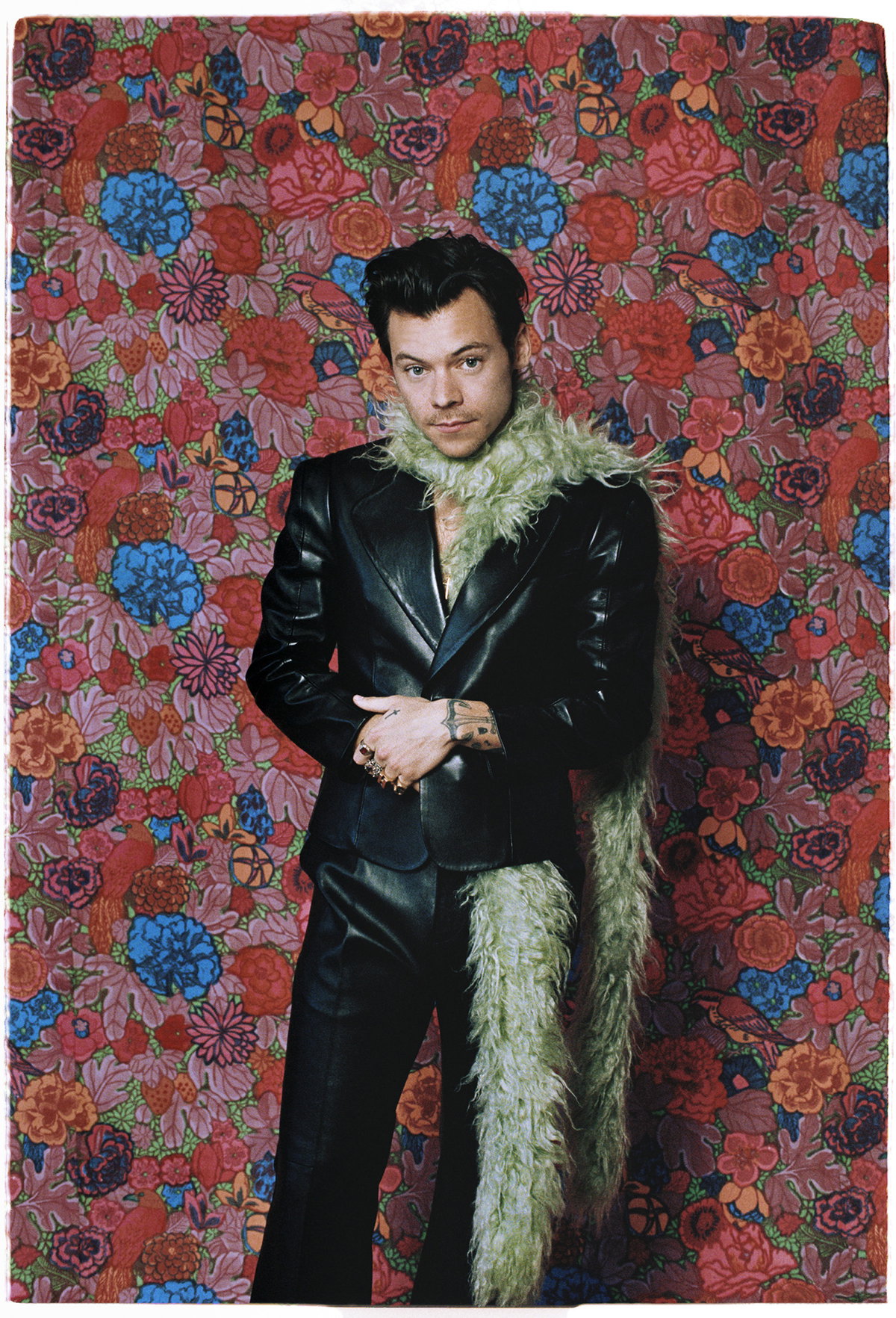 Harry Styles mit Federboa und schwarzem Anzug vor einer Blumenwand