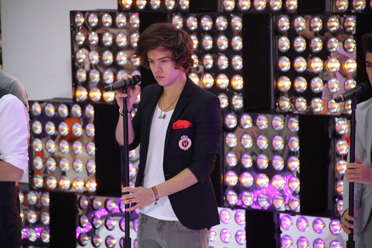 Harry Styles steht auf der Bühne und guckt ernst mit der Hand am Mikro 2012