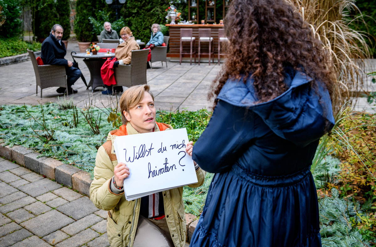 Sturm der Liebe Gerry kniet vor Shirin auf dem Boden und hält ein Schild mit den Worten "Willst du mich heiraten?" vor sich