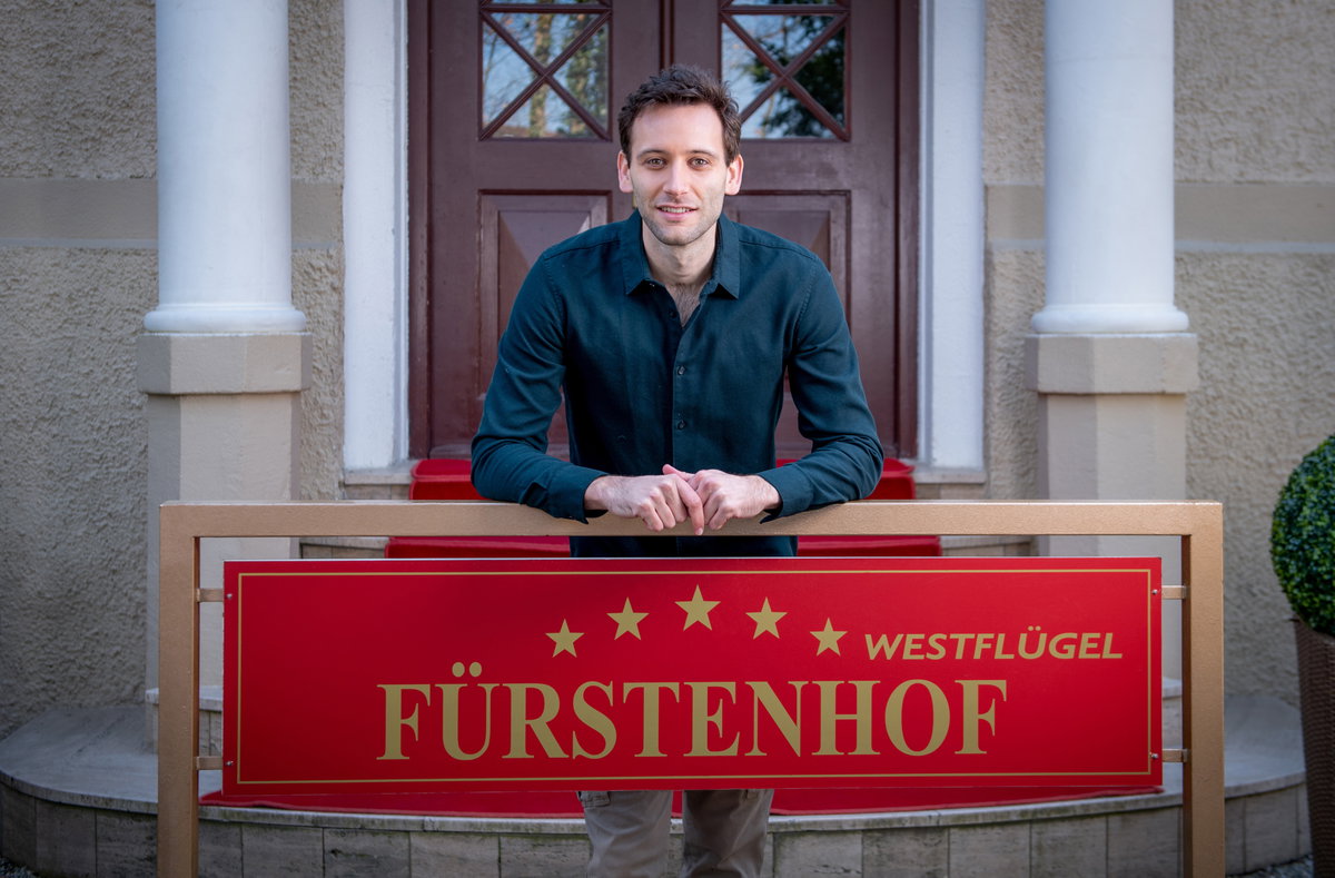 Sturm der Liebe Julian Specht lehnt an einem Fürstenhof-Schild vor dem Hotel