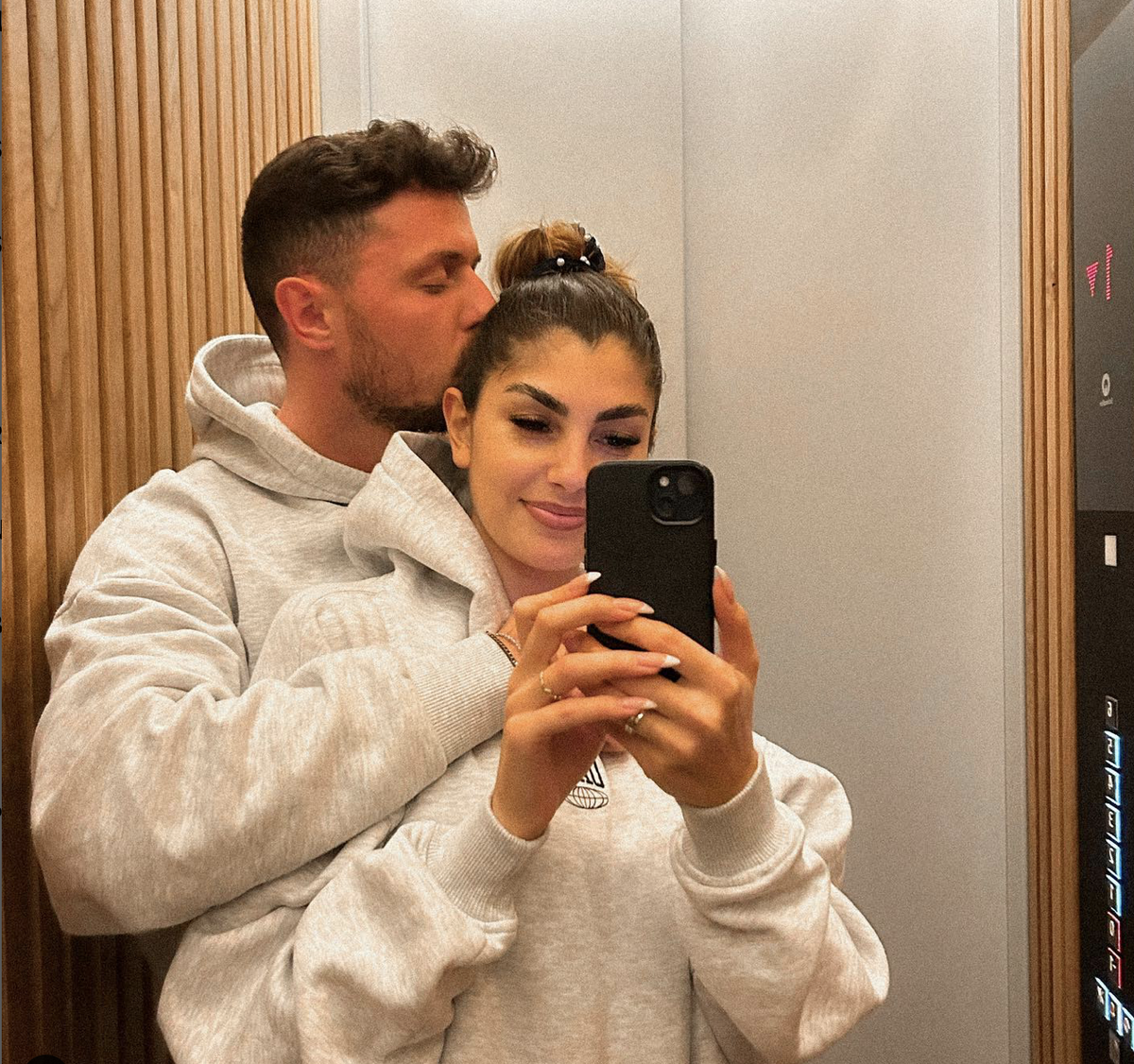 Yeliz Koc wird von hinten von Jannik Kontalis umarmt und bekommt einen Kuss auf die Wange während sie ein Selfie im Spiegel macht