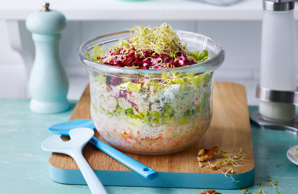Schichtsalat mit Feta-Dressing in einer Glasschale auf einem Holzbrett mit blauem Besteck daneben