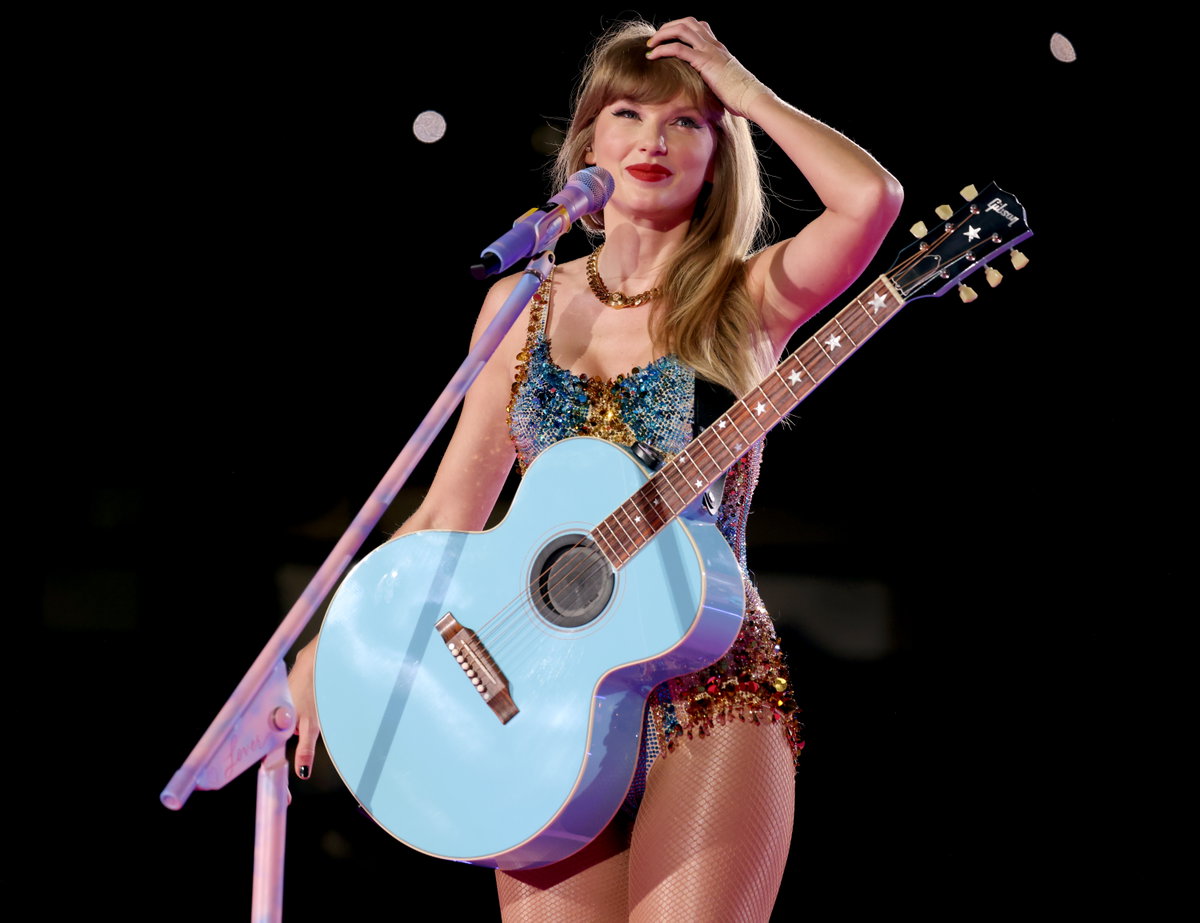 Taylor Swift mit Gitarre um den Bauch lächelt und fährt sich durch die Haare