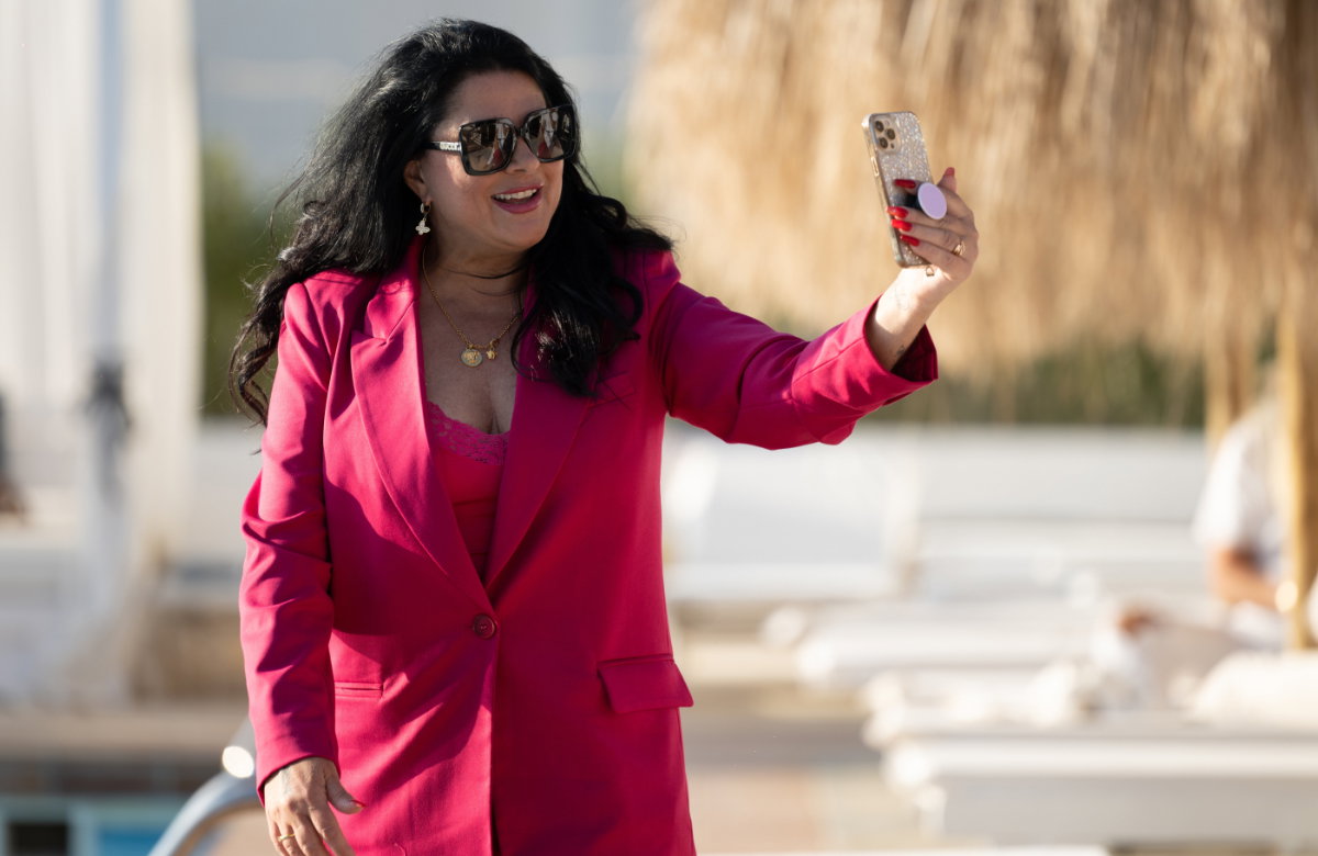 Iris Klein im pinken Anzug lacht und trägt eine Sonnenbrille, während sie mit Handy posiert