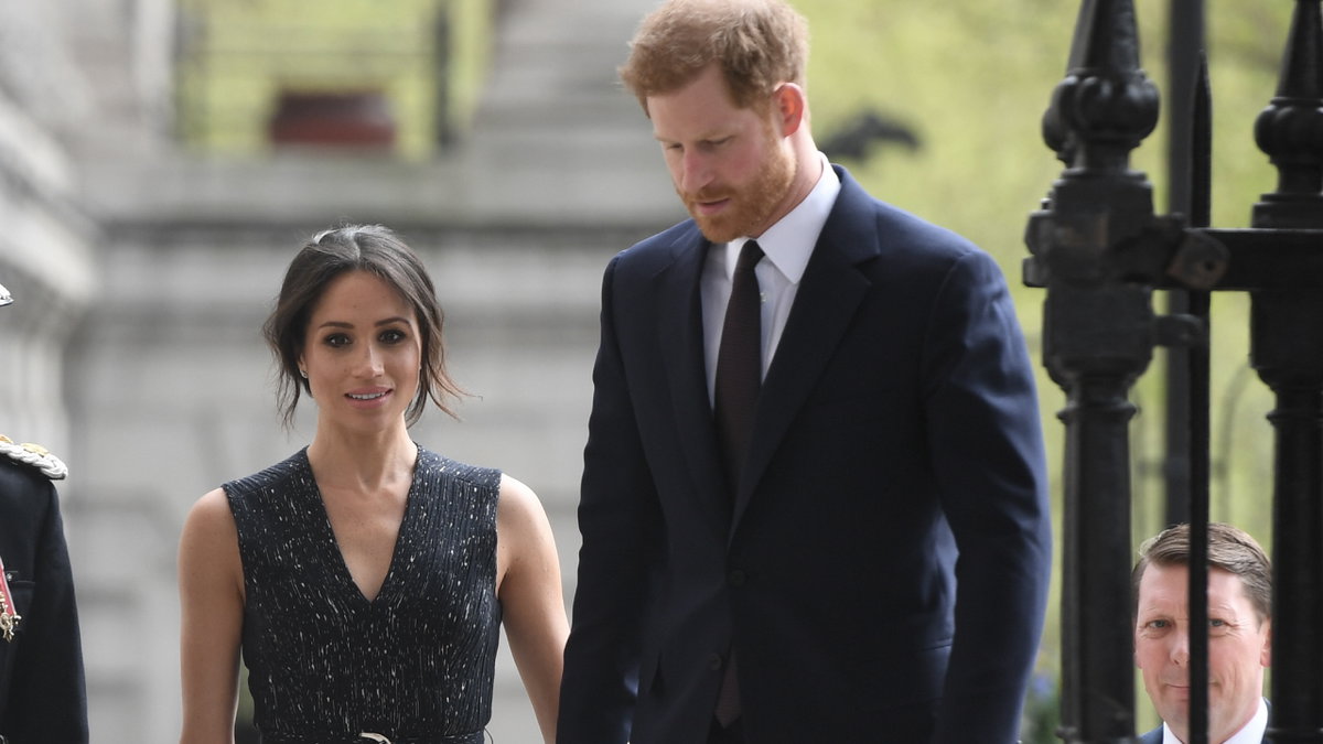 Prinz Harry sieht zu Boden, Herzogin Meghan läuft hinter ihm