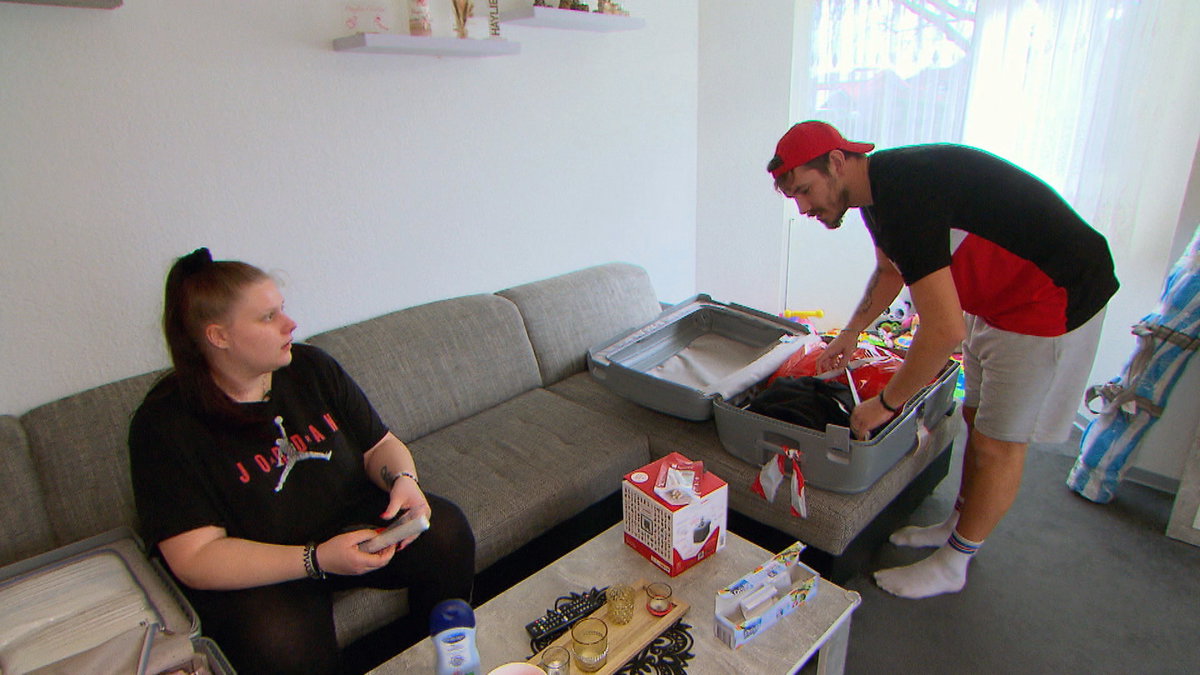 Lavinia Wollny sitzt auf der Couch, Tim Katzenbauer packt einen Koffer