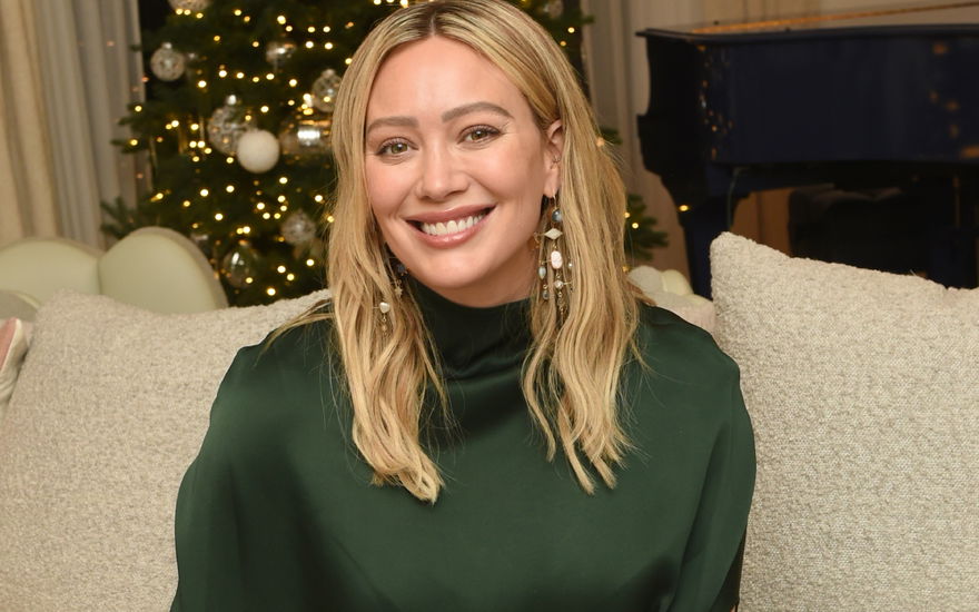 Hilary Duff sitzt lächelnd auf der Couch, im Hintergrund steht ein Weihnachtsbaum