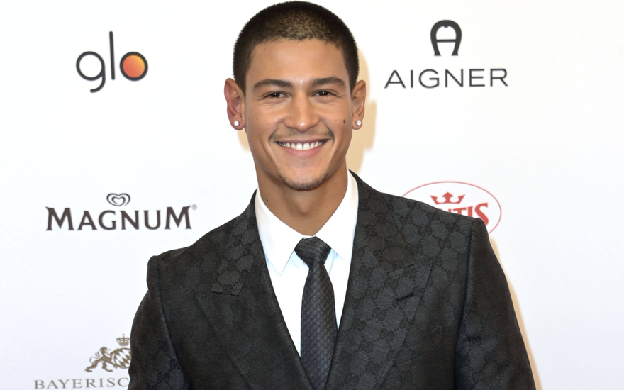 Emilio Sakraya im schwarzen Anzug grinst vor einer weißen Wand beim Deutschen Filmball 2024