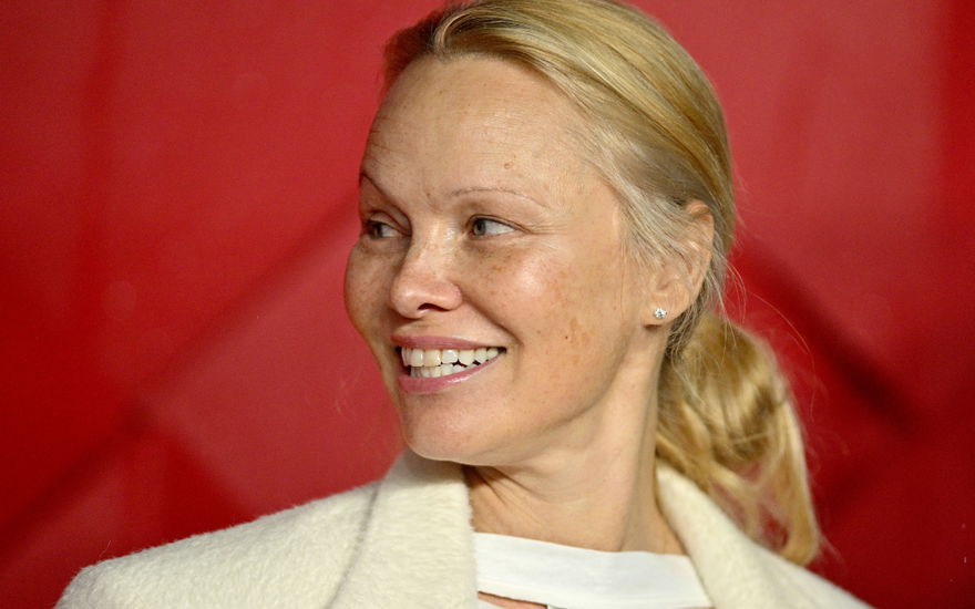Pamela Anderson guckt zur seite und lächelt vor rotem Hintergrund