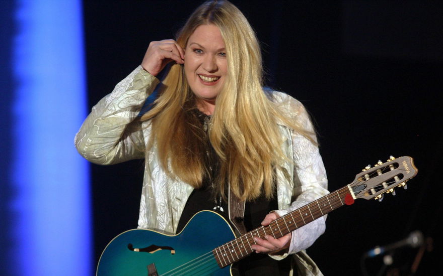 Juliane Wedding steht mit Gitarre auf der Bühne und fasst sich lächelnd ans Ohr