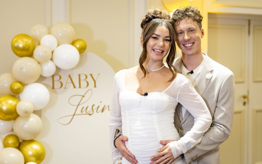 Valentin Lusin steht hinter Renata Lusin in einem weißen Kleid und umarmt sie bei ihrer Babyparty