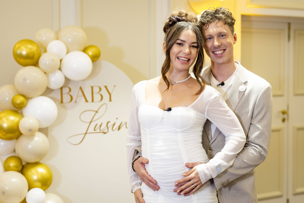 Valentin Lusin steht hinter Renata Lusin in einem weißen Kleid und umarmt sie bei ihrer Babyparty