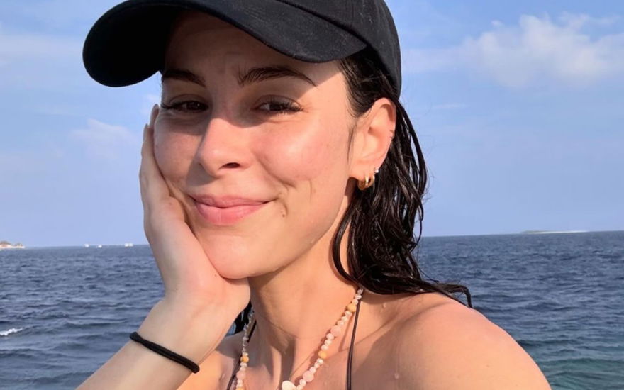 Lena Meyer-Landrut sitzt lächelnd mit Cap und Bikini am Meer