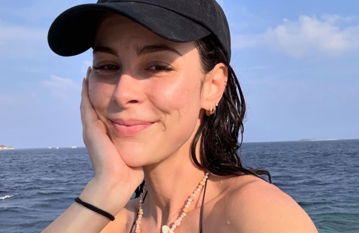Lena Meyer-Landrut sitzt lächelnd mit Cap und Bikini am Meer