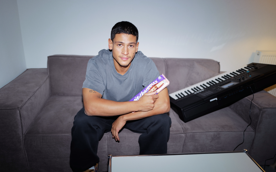 Emilio Sakraya sitzt auf einer Couch mit einer Tafel Milka in der Hand und einem Keyboard neben sich