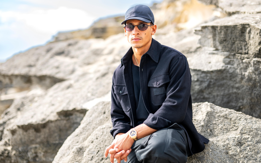 Emilio Sakraya mit Sonnenbrille auf sitzt auf einem Stein am Strand