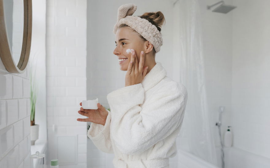 Junge Frau lächelt in Spiegel und benutzt Anti-Aging-Creme