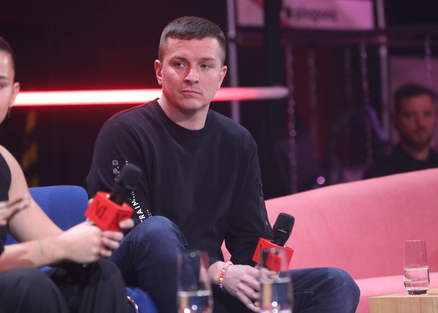 Aaron Troschke sitzt auf einem Sofa mit einem Mikrofon in der Hand und schaut ernst