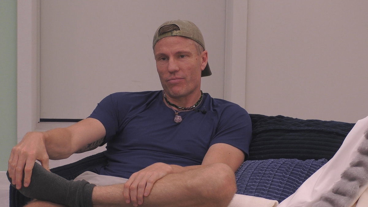 Christian Lell sitzt bei "Promi Big Brother" mit Cappy auf dem Sofa und grinst