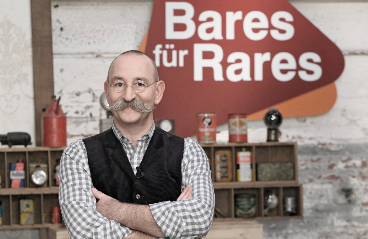 Horst Lichter mit verschränkten Armen vor dem "Bares für Rares"-Logo